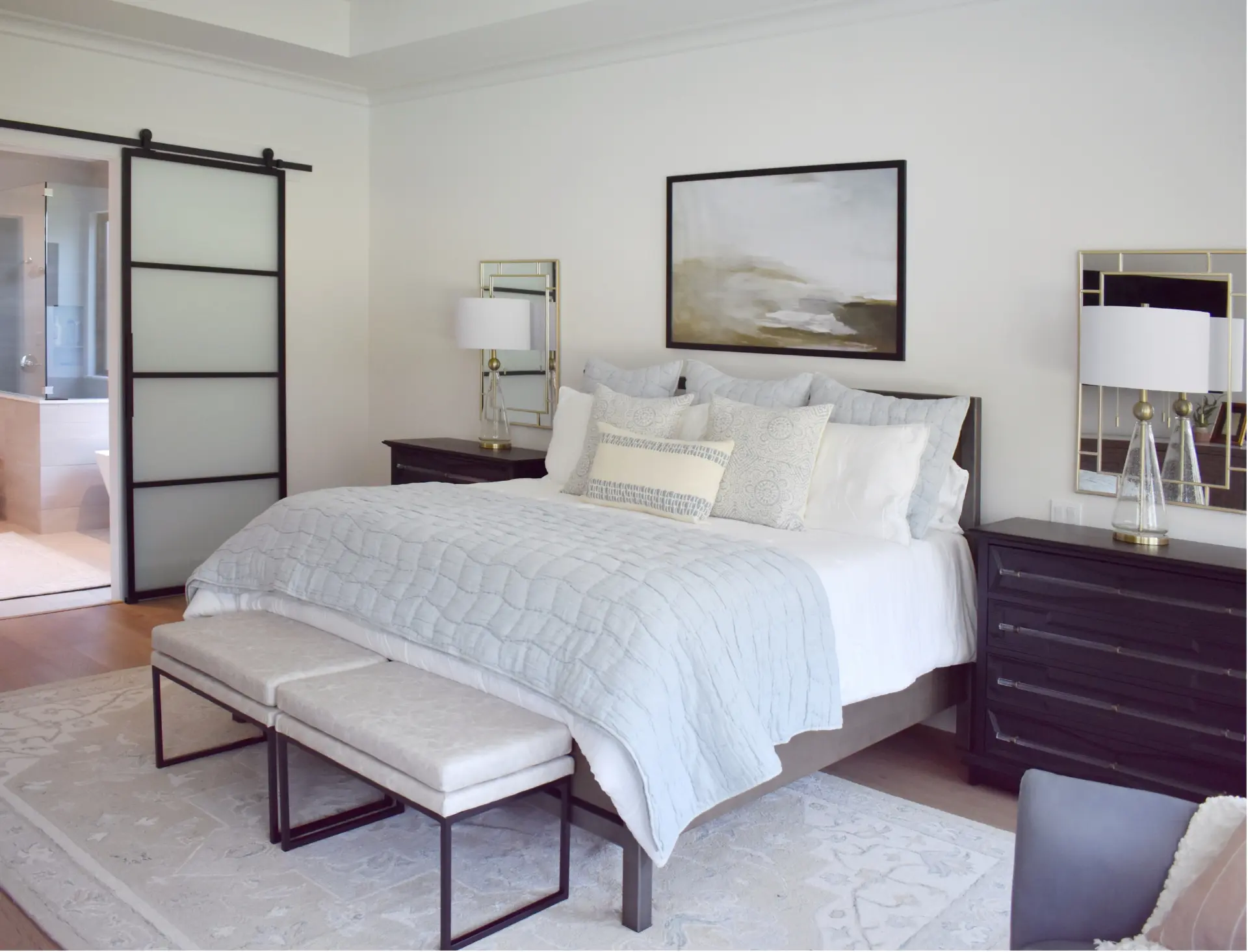 Finished master bedroom with white bedding, two benches at the foot of the bed, and a sliding barn door to the en-suite bath